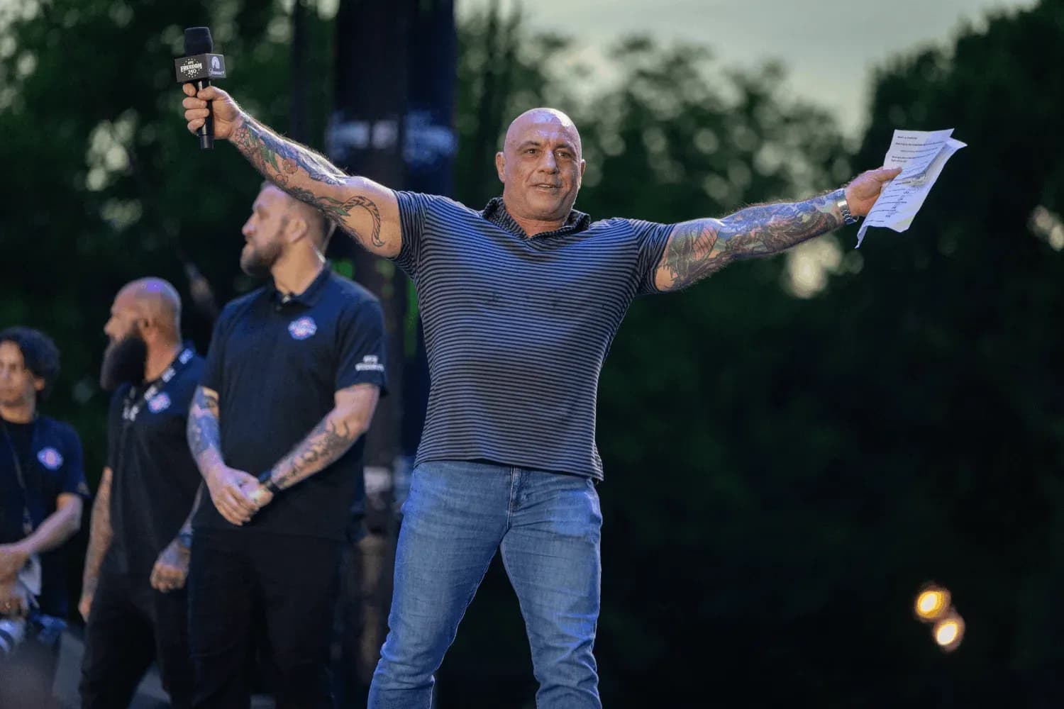 Joe Rogan spreads his arms wide on stage while hosting a UFC event.